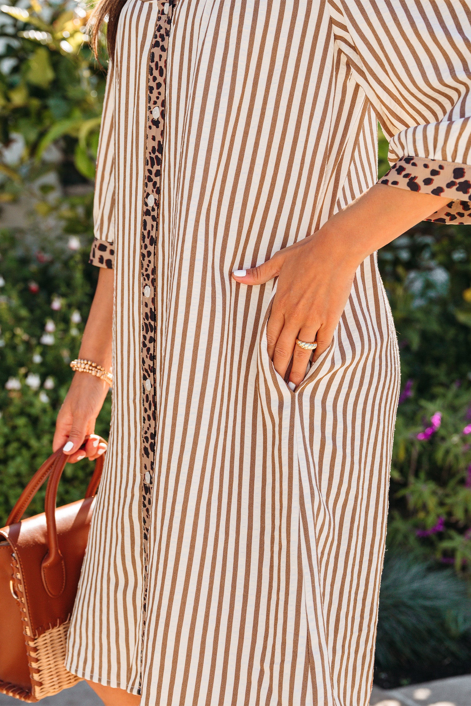 Mulher vestindo Khaki Stripe Square Button Leopard Trim Loose Bracelet Sleeve Shirt Dress com padrão de listras kaki e detalhes em estampado de leopardo, segurando uma bolsa de mão, em ambiente ao ar livre com vegetação ao fundo, transmitindo um estilo casual e elegante.