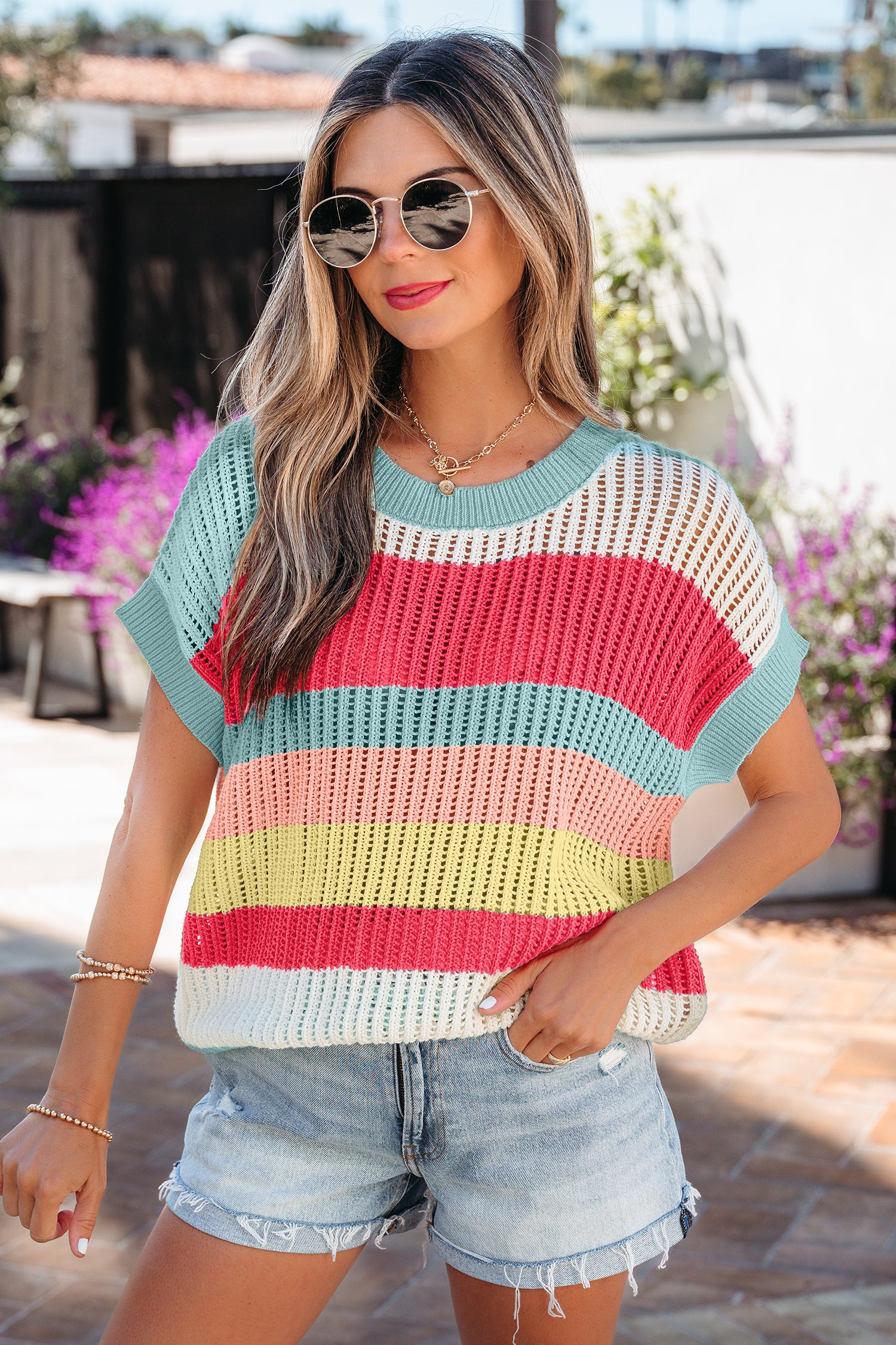Mulher sorridente usando Green Stripe Color Block Hollow Out Knit Short Sleeve Sweater colorido com shorts jeans, refletindo estilo casual e descontraído em ambiente externo iluminado.