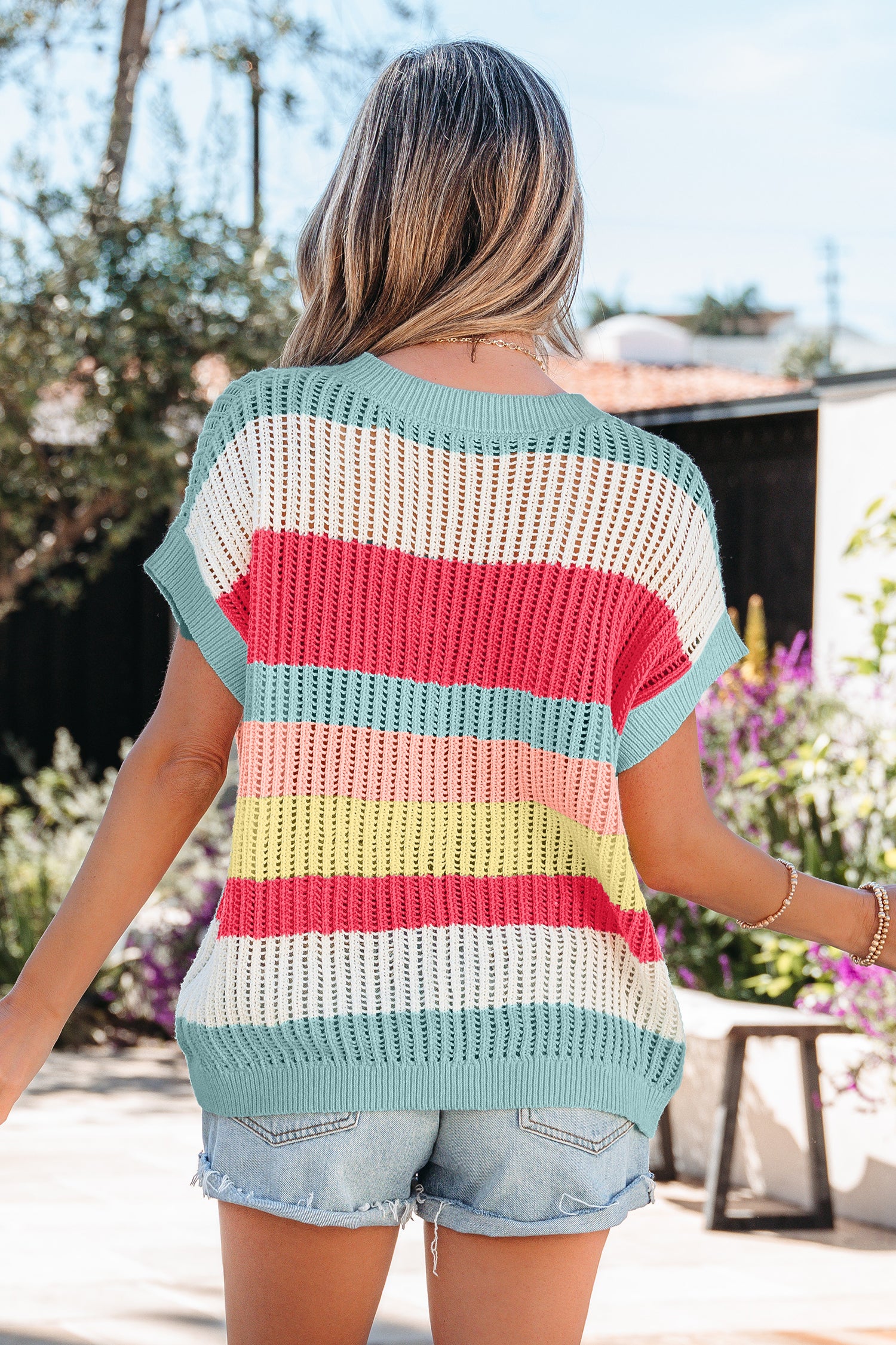 Mulher vista por trás usando Green Stripe Color Block Hollow Out Knit Short Sleeve Sweater com listras coloridas e calções de ganga, em ambiente exterior ensolarado, transmitindo estilo casual e fresco.