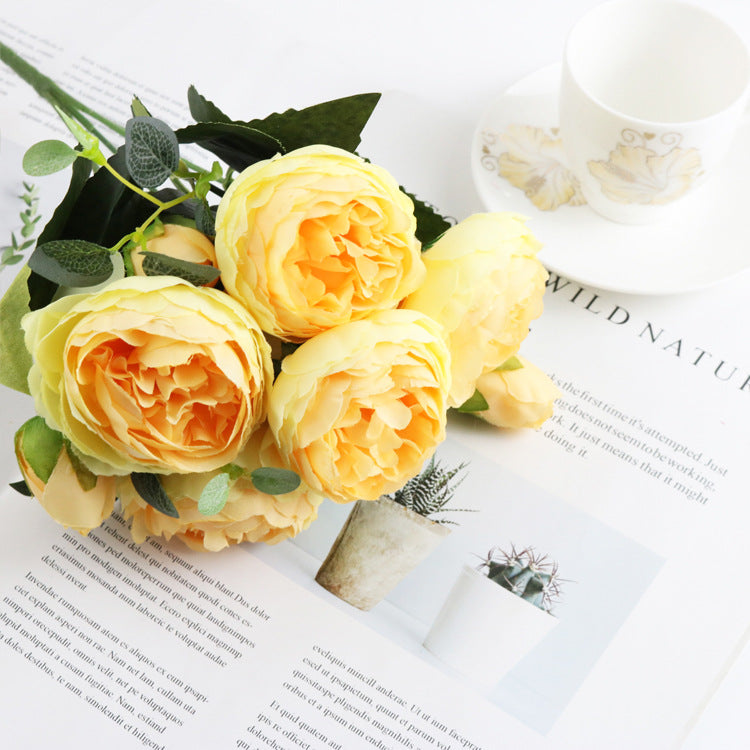 Arranjo delicado de flores amarelas com folhas verdes sobre revista, acompanhando a descrição detalhada de "5 Small Peonies Korean Small Bouquets 5 Philips Rose Simulation Flowers Household Wedding Rose Bouquets", realçando suplementos premium naturais para energia, vitalidade e saúde cognitiva, desenvolvidos para o mercado português.