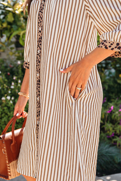 Mulher vestindo Khaki Stripe Square Button Leopard Trim Loose Bracelet Sleeve Shirt Dress com padrão de listras kaki e detalhes em estampado de leopardo, segurando uma bolsa de mão, em ambiente ao ar livre com vegetação ao fundo, transmitindo um estilo casual e elegante.