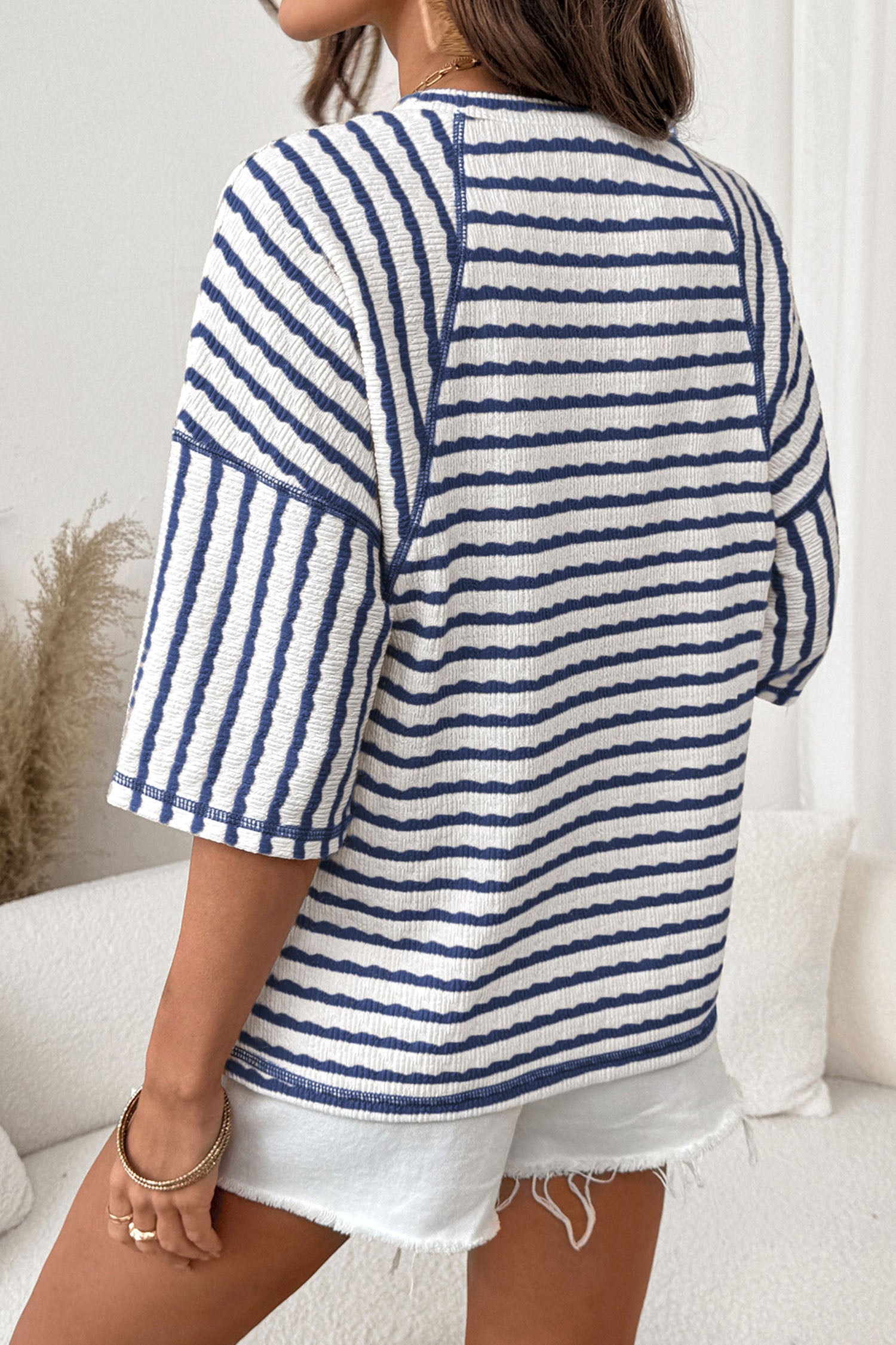 Mulher vista de costas usando Apricot Stripe Textured Knit Split Hem Half Sleeve Loose Top com padrão listrado azul e branco, combinando com shorts brancos em ambiente claro e acolhedor, suplemento premium naturais para energia, vitalidade e saúde cognitiva.