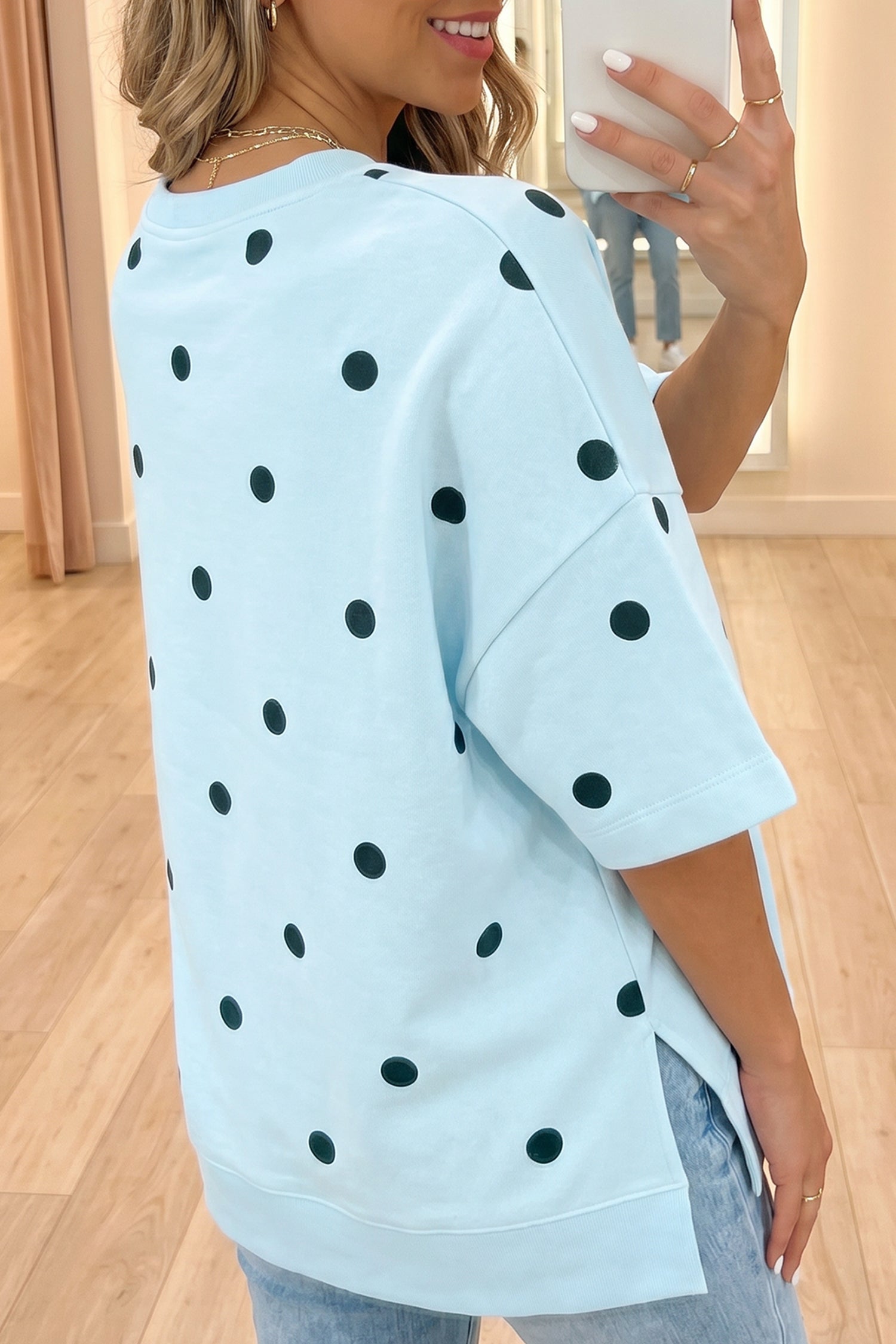 Mulher vestindo Yellow Polka Dot Print Crossed Seam Detail Oversize Tee azul clara com bolinhas pretas, em ambiente interno moderno com chão de madeira clara.
