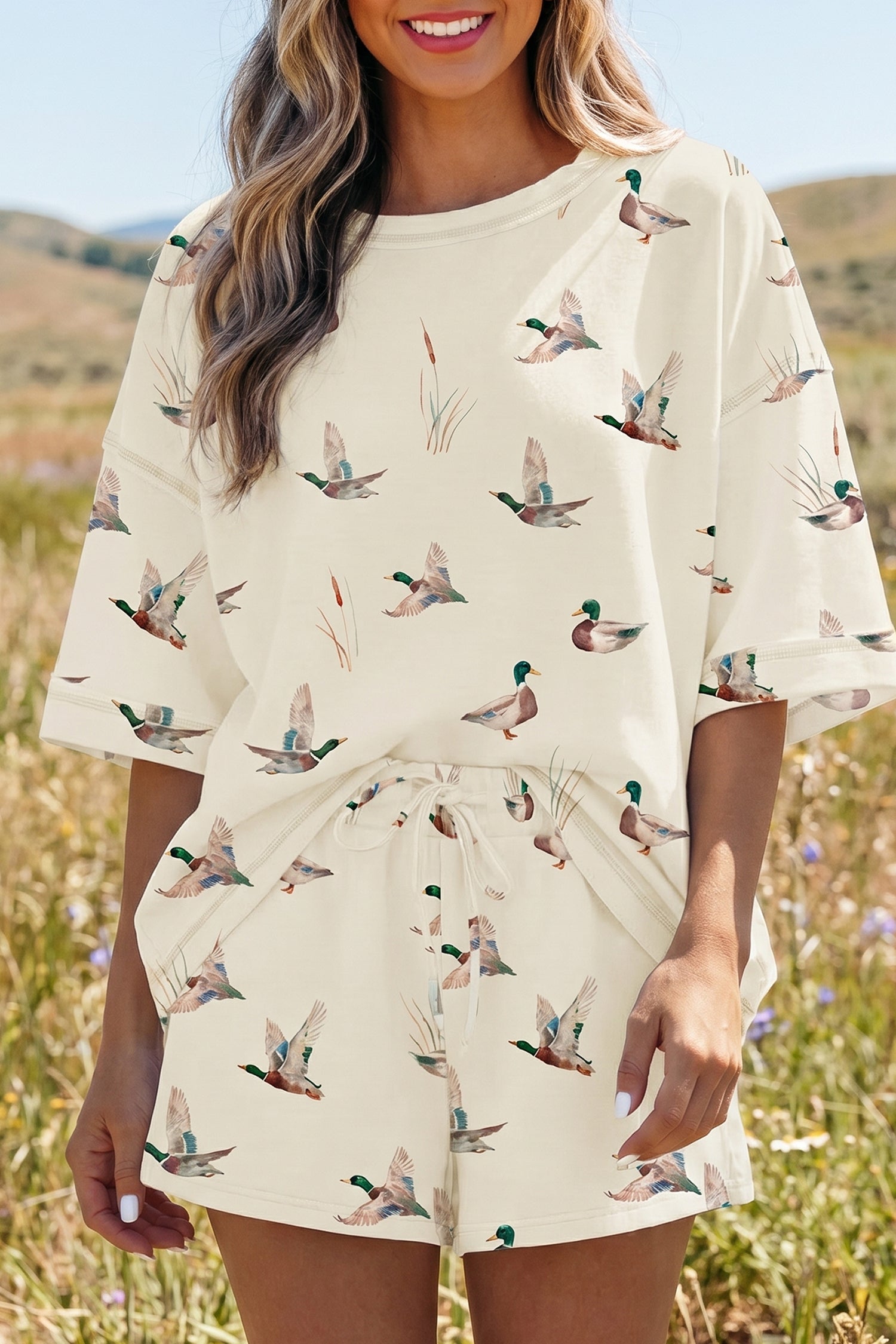 Mulher sorridente usando Beige Mallard Duck Print Oversized Tee Drawstring Short Set em tom bege com estampa de patos, transmitindo conforto e estilo natural ao ar livre.