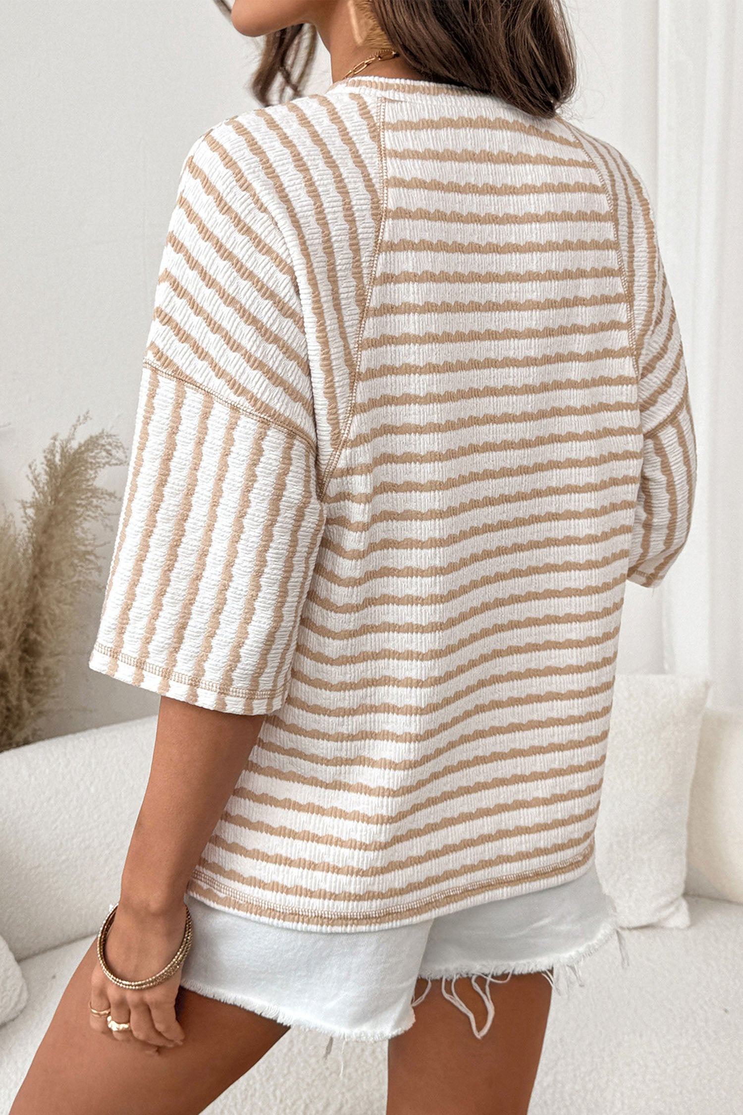Mulher vista de costas usando Apricot Stripe Textured Knit Split Hem Half Sleeve Loose Top com riscas texturizadas em bege e branco, combinada com calções brancos rasgados, em ambiente claro e tranquilo.