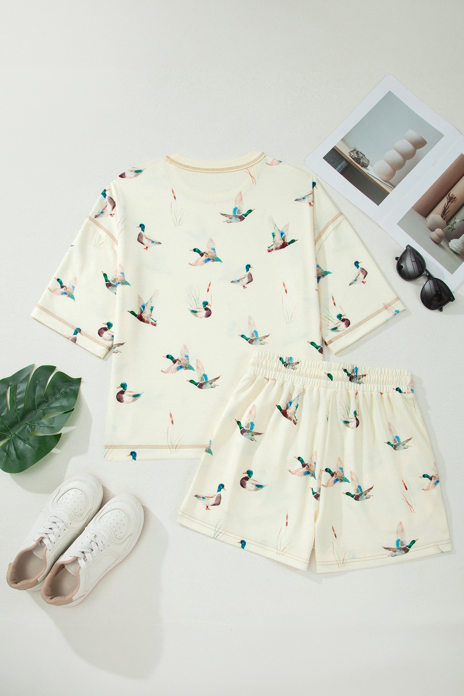 Conjunto Beige Mallard Duck Print Oversized Tee Drawstring Short Set com estampa de patos mallard em bege, acompanhado de sapatilhas brancas, óculos de sol e revistas, ideal para um look confortável e casual, remetendo a suplementos premium naturais para energia, vitalidade e saúde cognitiva, desenvolvidos para o mercado português.