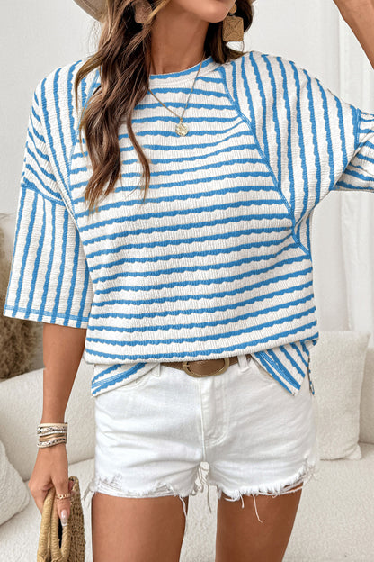 Mulher vestindo Apricot Stripe Textured Knit Split Hem Half Sleeve Loose Top em tons claros com listras azuis, combinando com shorts brancos, transmitindo estilo casual e conforto.